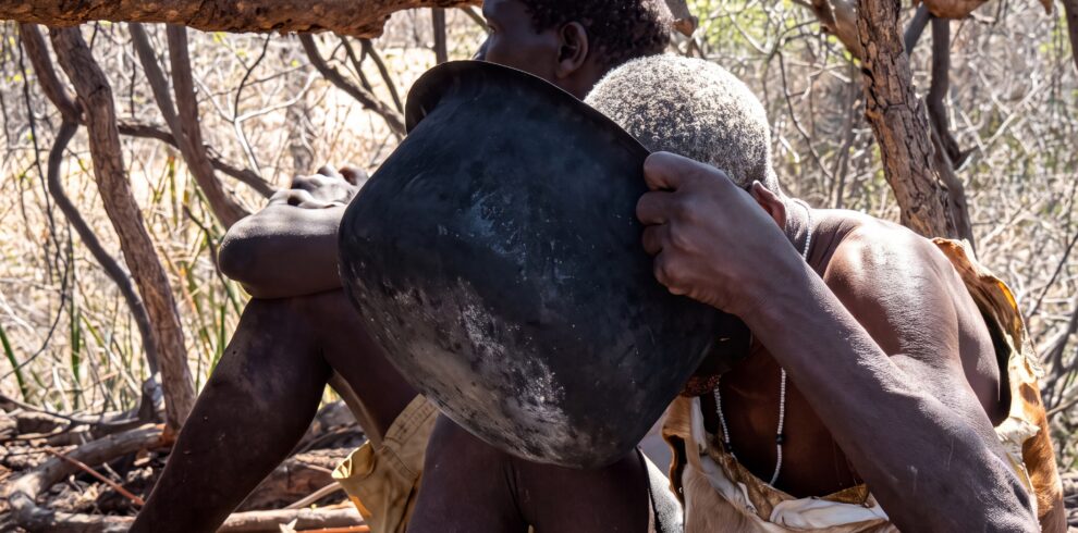 Two Hadzabe elders seated together beneath the shade of a tree near Lake Eyasi, sharing a quiet moment as one drinks from a metal cooking pot, reflecting the simplicity and continuity of daily life.